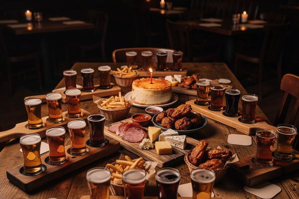 A long wooden pub table covered with beer flights, shared platters, and a small cake during a party